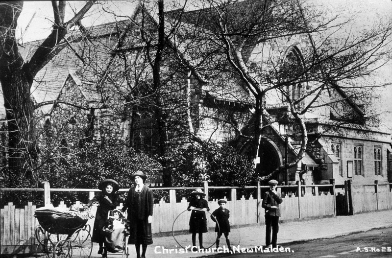 Photo of Christ Church taken in the 1920s, partly obscured by a tree; in the foreground is a young mother with perambulator, with several children (two with hoops)