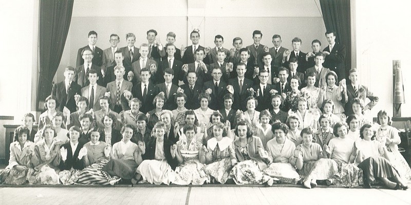 Group photo of Kings Own taken in the Roberts era with a stage as backdrop, girls seated in front rows and boys standing in the back rows