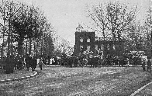Photo of Alfred Streeter funeral, horse drawn hearse bedecked with flowers, with Malden College in the background